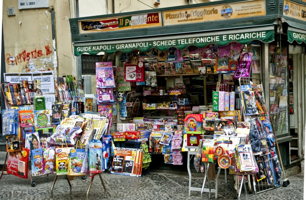 The space in front of a corner news stand is filled with racks of colorful magazines and toys for children.
