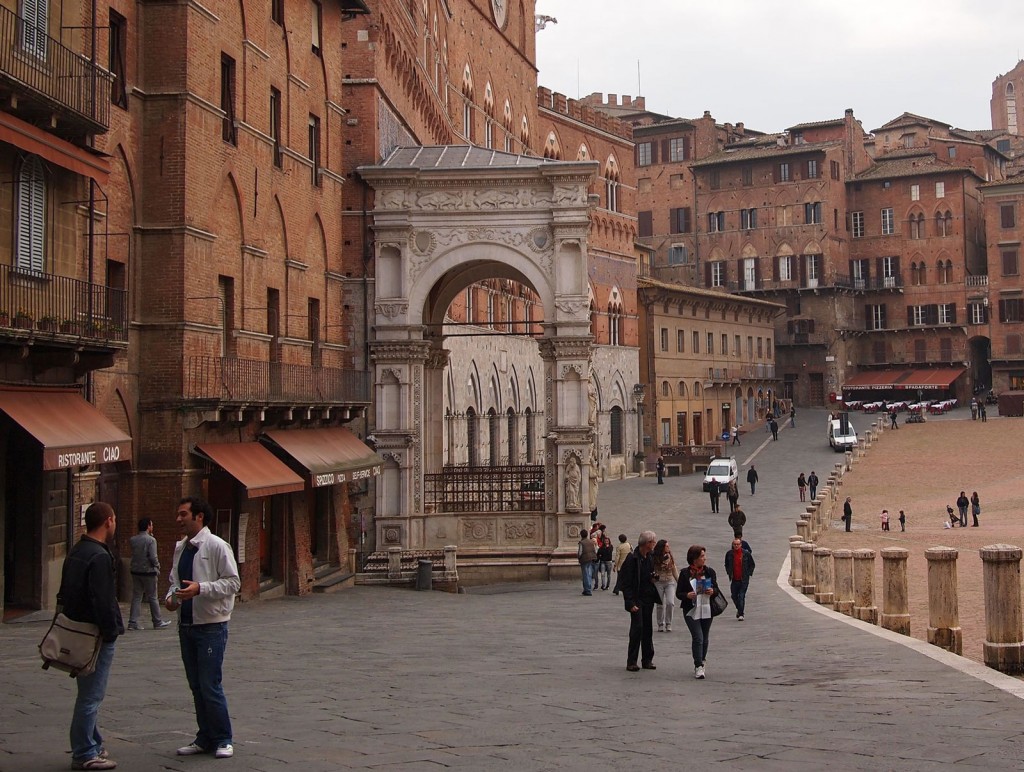 Tourists and pedestrians stroll along the edge of a plaza lined with multi-story brick buildings, some with carved stone facades.