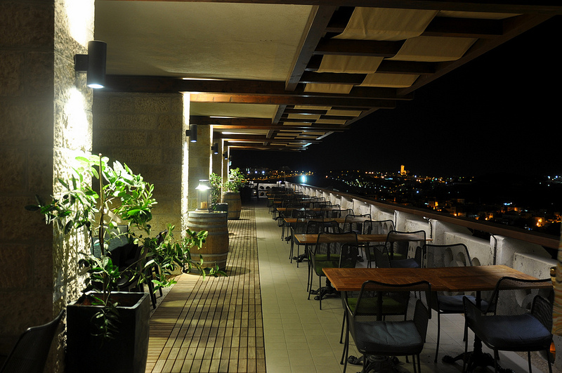 Tables line up against the edge of a balcony with a view of the city lights spreading to the horizon.