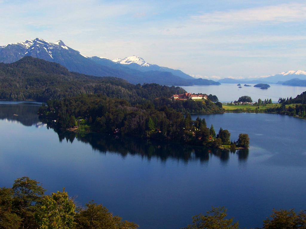 Snow-crested mountains are visible in the distance while the heavily forest shore reflects in deep blue lakewater.