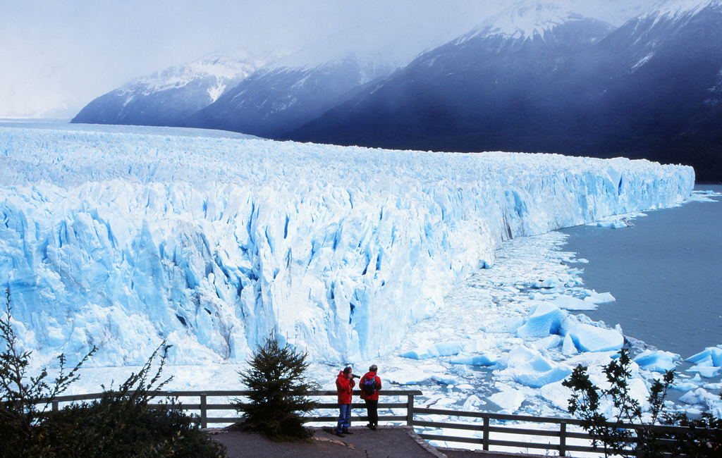 The edge of the glacier sheds into the water while snow-capped mountains rise up across the lake.