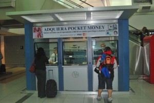 Travelers stand at a small kiosk to exchange money with a teller.