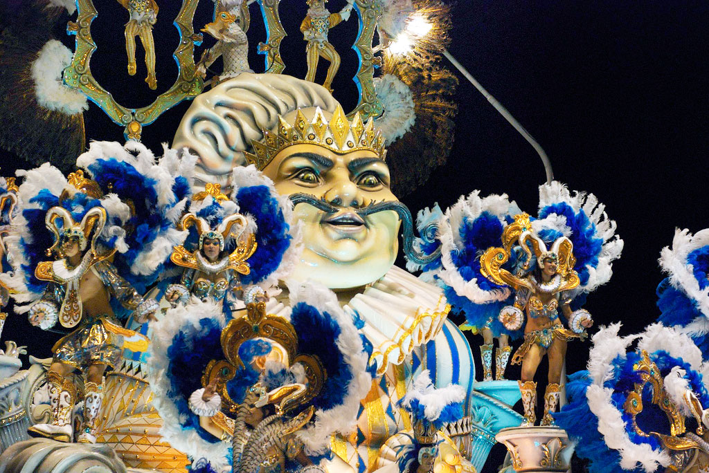 Dancers in feathered costumes stand on gilded pillars on an elaborate float featuring a large carved face.