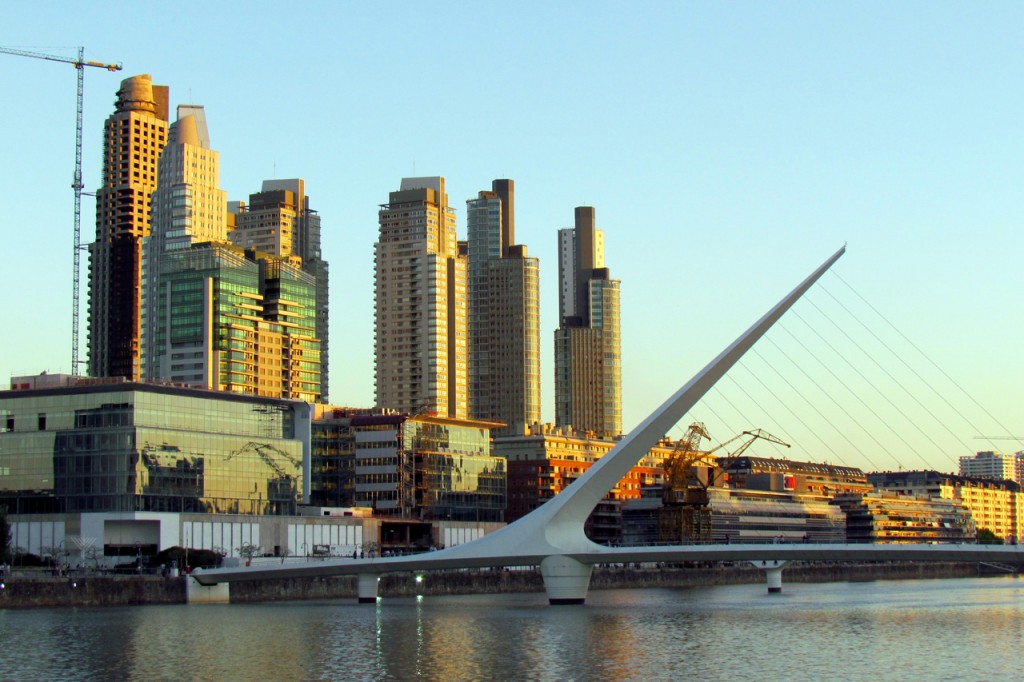 View of the waterfront with a elegant white footbridge crossing the water and skyscrapers in the background.