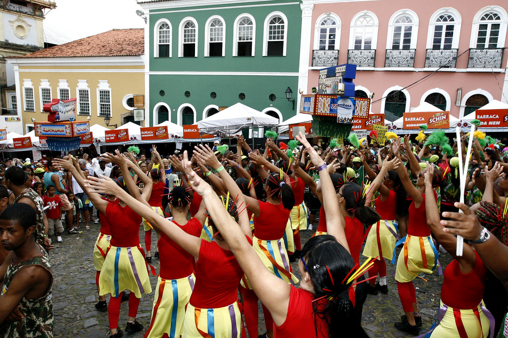 In a cobblestoned square surrounded by colorful colonial buildings, a group of dancers in red shirts and yellow ribboned skirts dance with their arms raised.