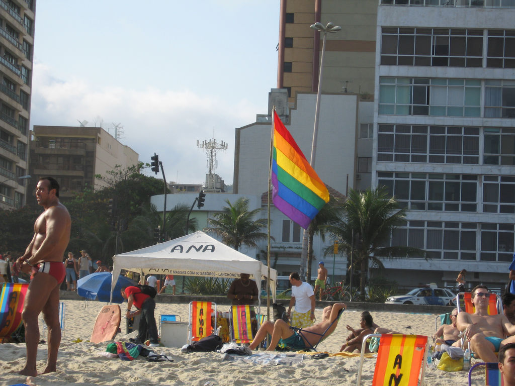 Beachgoers near a tent with a rainbow flag and beach chairs on the white sand of Ipanema beach.