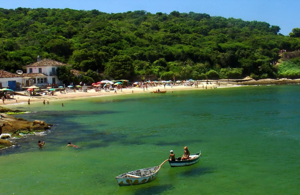 Rowboats float in the clear green waters off a busy beach.