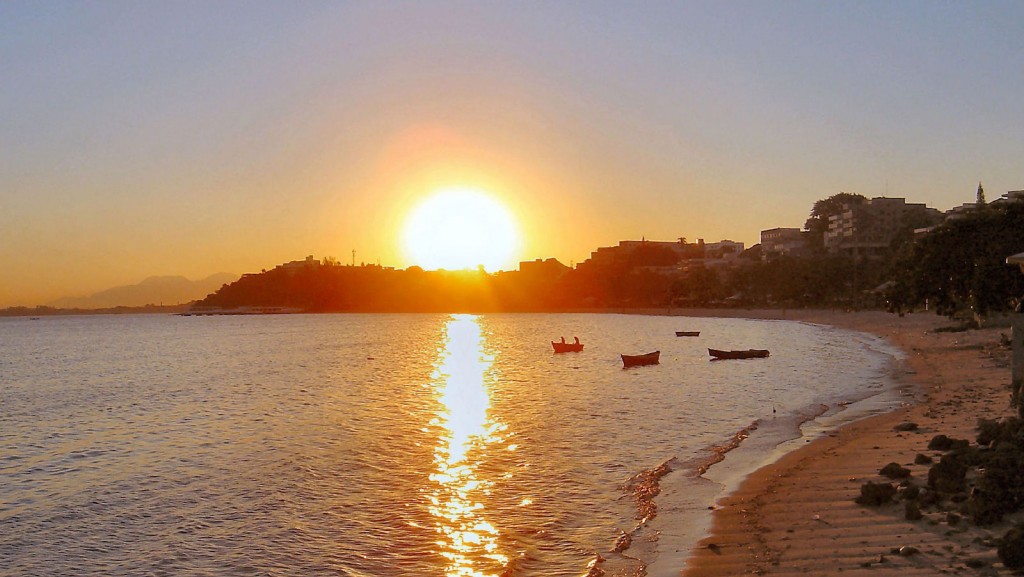 The sun sets on a Rio beach with small two-person boats floating int he calm water.