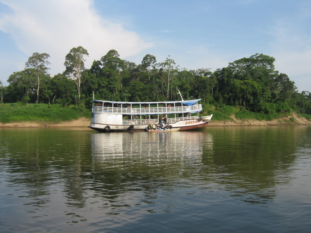 A two-level river boat with a smaller boat alongside floats on glassy river water at the verdant shore.