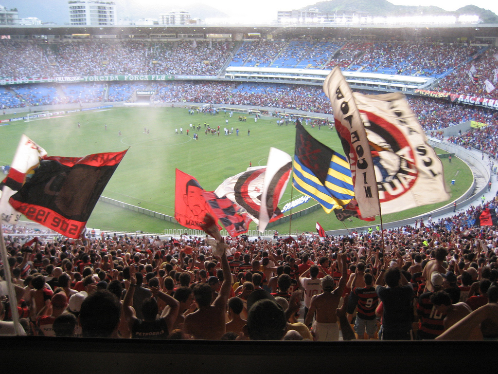 View from the soccer stadium stands where fans wave flags and smoke curls into the air.