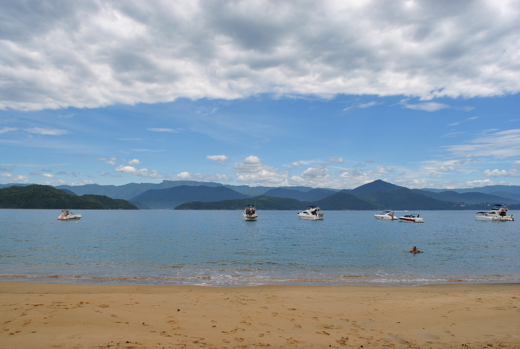 View of calm ocean water from a flat, golden sand beach with hills overlapping far in the distance.