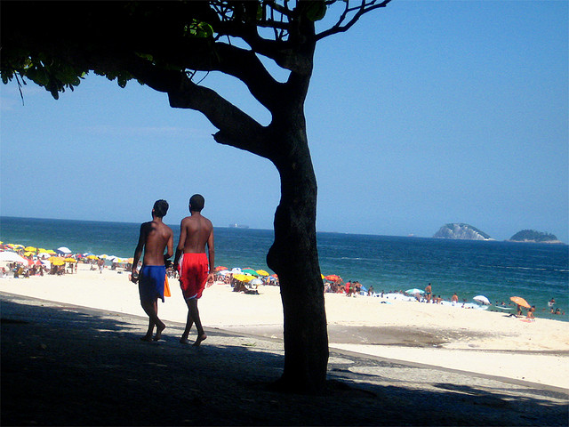 Two men pass under the shade of a tree high up on the shore of a white-sand beach.