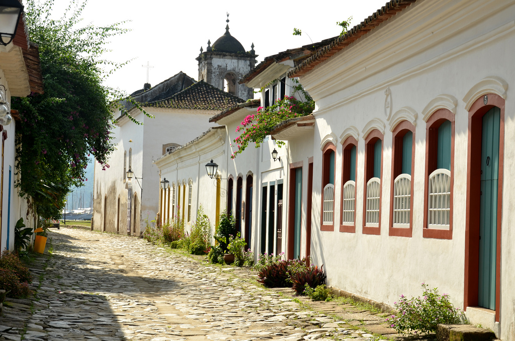 View down a cobblestoned street lined with white-washed colonial single story buildings and the view of the harbor in the distance.