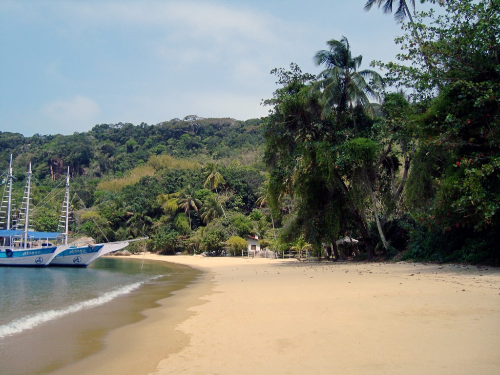 Two boats float offshore as gentle waves lap on the shore of a wide sandy beach with dense tropical foliage just beyond.