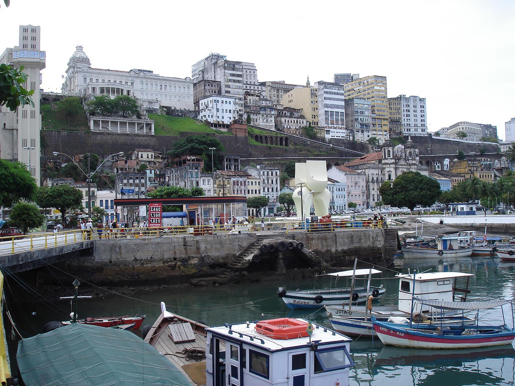 View of pedestrians walking along the waterfront as buildings stack up against a terraced hillside and boats cluster in the water.