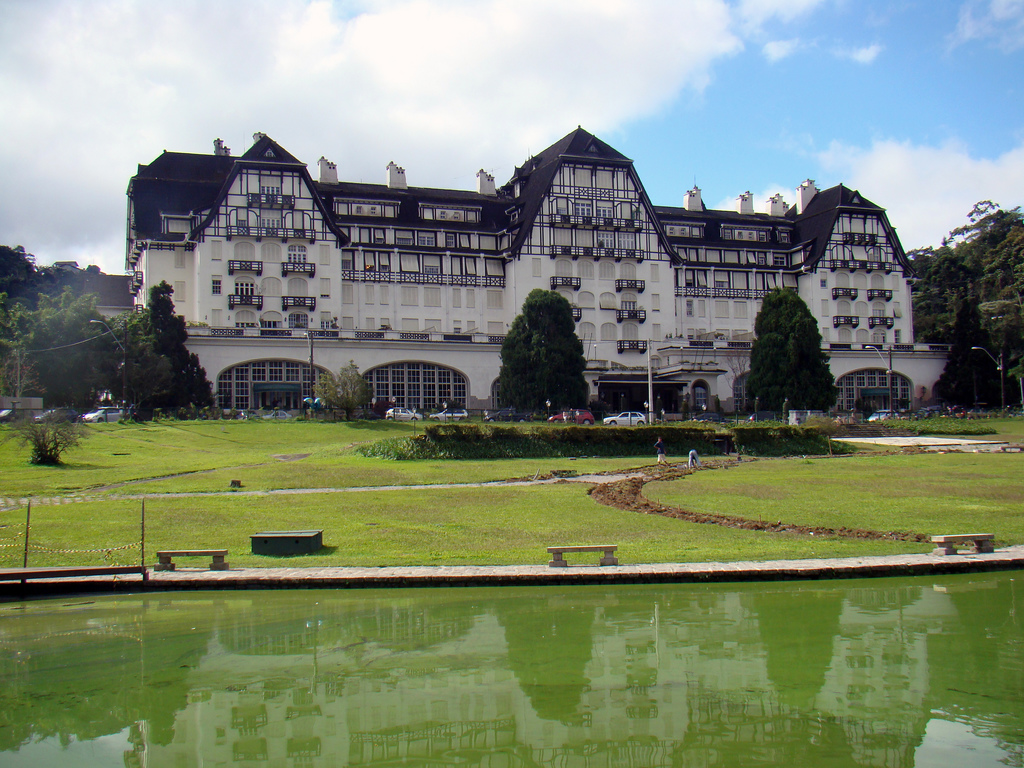 View of a multi-story Norman-style palace with a neatly trimmed lawn sloping down towards a reflecting pool.