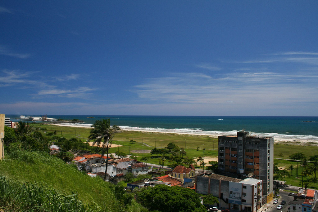 View atop a grassy hill of a small strip of buildings and a span of grass between them and a long stretch of beach.