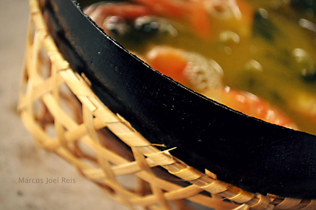 View of broth with visible pieces of shrimp in a cooking pan cradled in a woven basket.