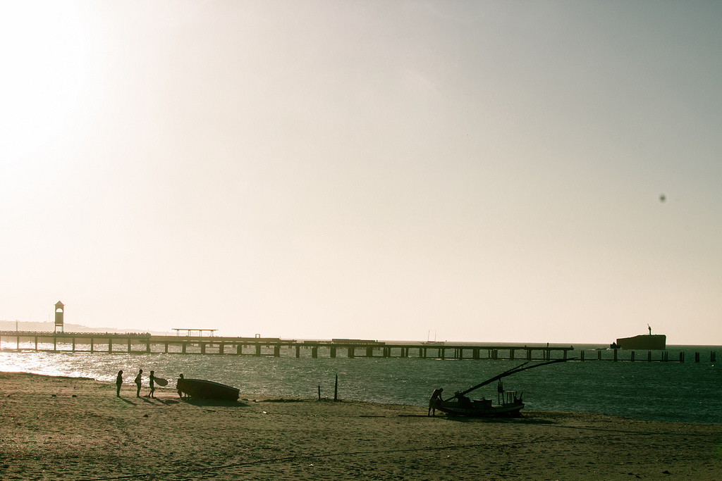 At dusk a couple boats are pulled up on to the sand with a long pier visible in the water.