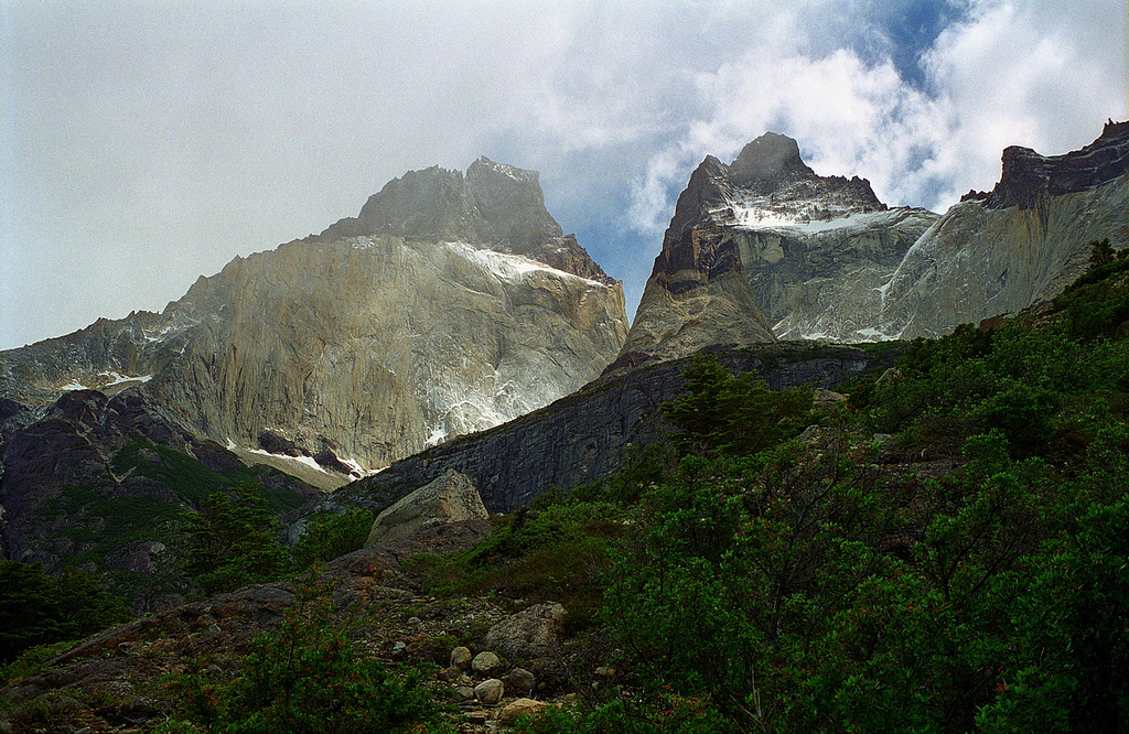 Viewed from a trail lined with scrub, raw stone dusted with snow rise up in peaks.