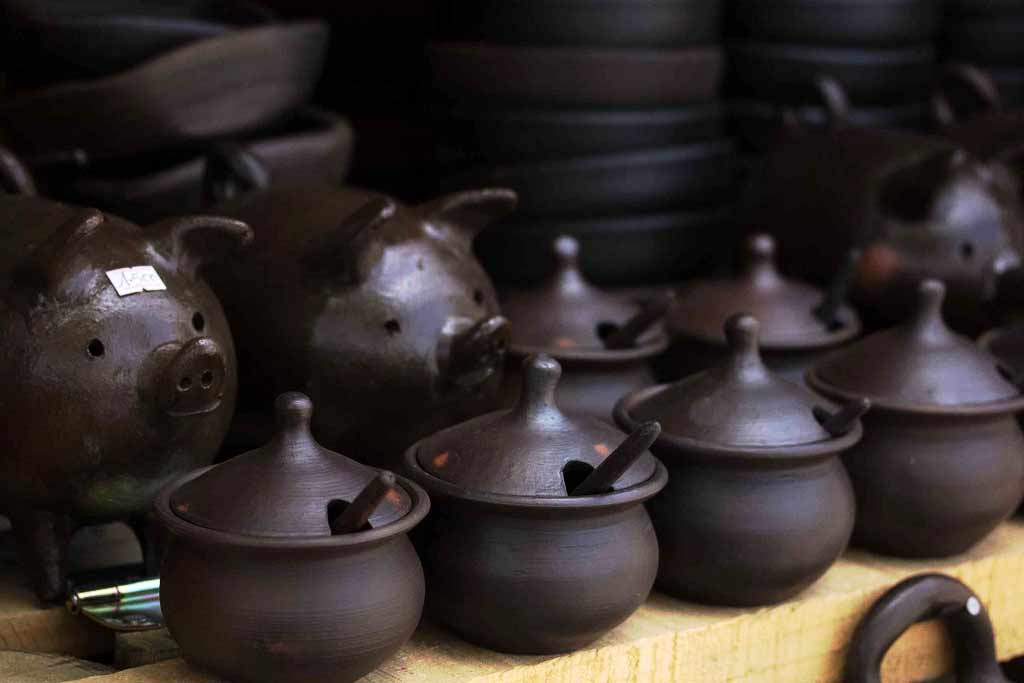 Rows of dark-glazed pottery for sale: round baking dishes, pots with spoons, and ceramic pigs.