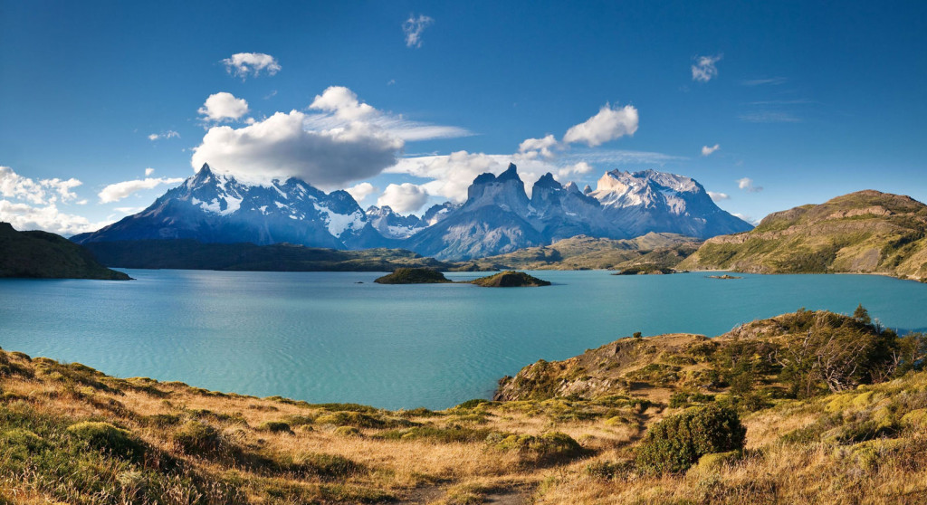 Striking blue glacial mountains rise up beyond an aquamarine lake with the stark contrast of dry grasses at the near shore.