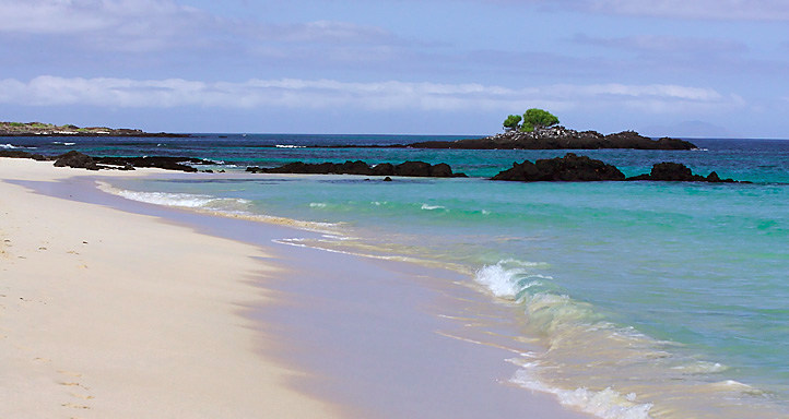 Soft waves lap on a light sand beach with rocks jutting out of the water at a distance.