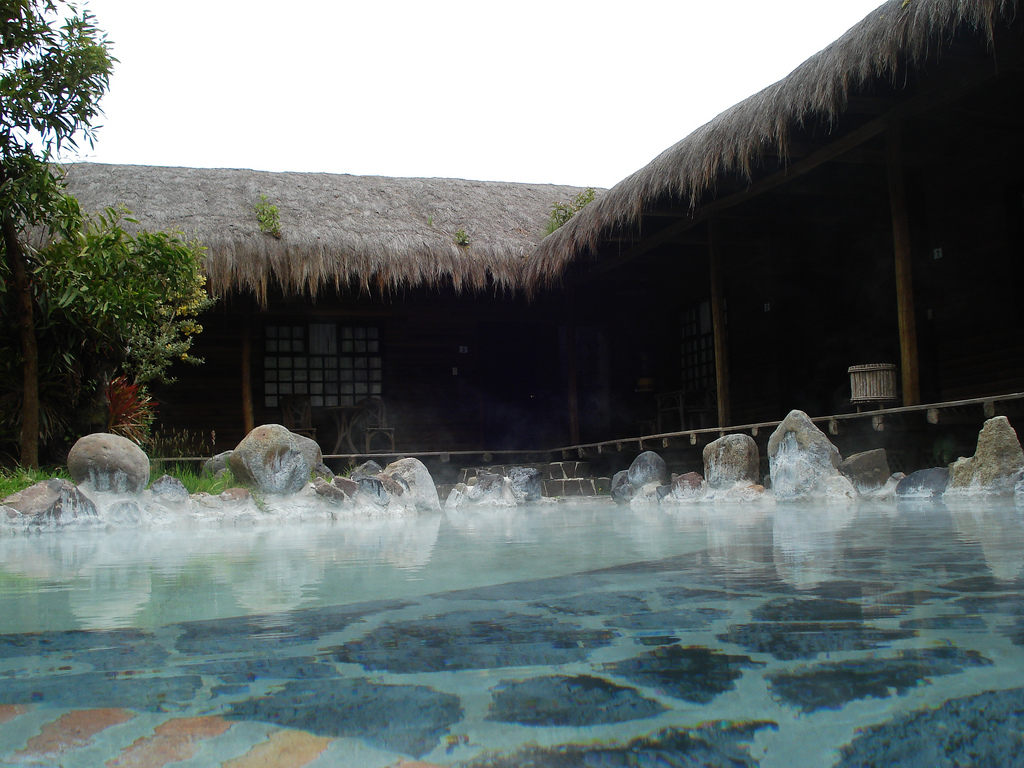 A shallow pool of steaming water ringed by cabins with thatched roofs.