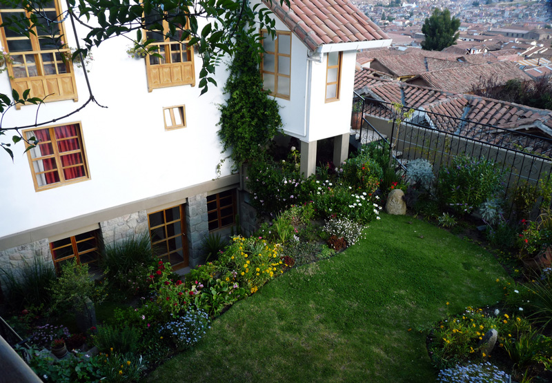 A two-story building with whitewashed walls and tile roof rises above a charmingly manicured backyard garden.