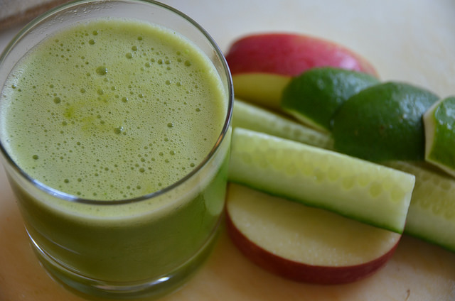 A glass of green juice next to sliced fruits and vegetables.