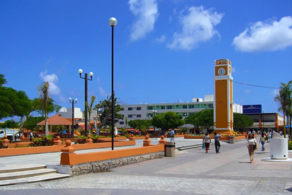 Pedestrians walk through a plaza with an orange and white stucco clocktower visible at the far end.