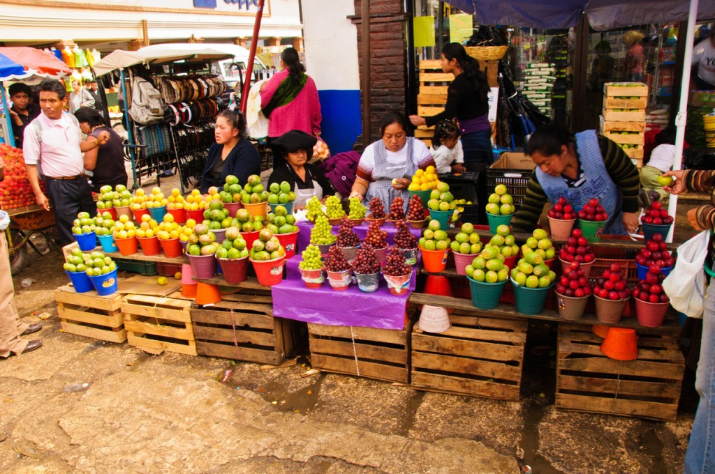 Small buckets filled with fruit stacked pyramidally are sold by women in a busy market.