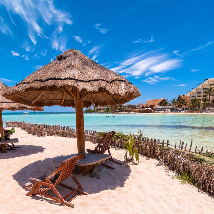 A palm umbrella and wooden chairs grace the beach near a shallow shore.