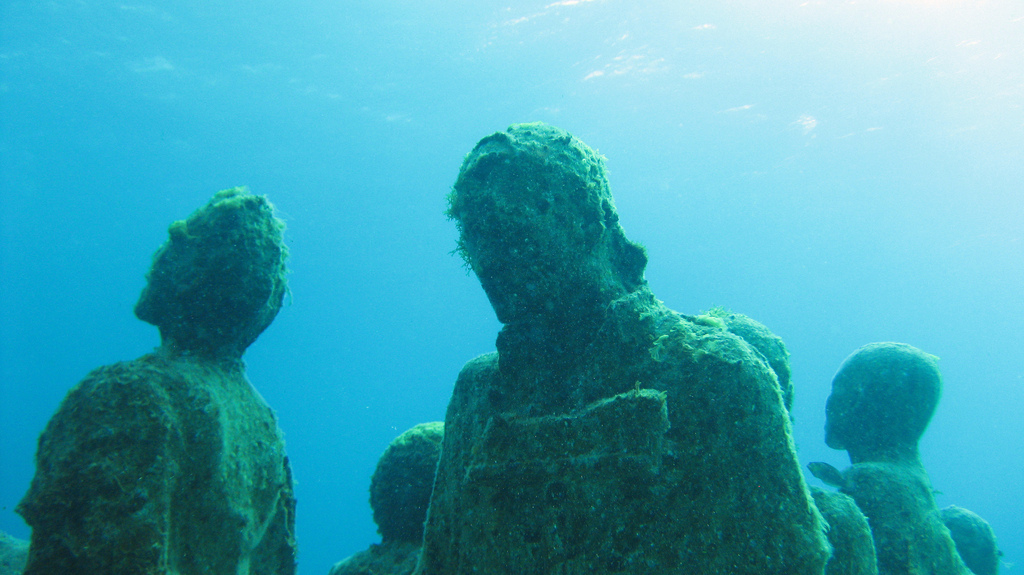 Sculpted statues underwater are covered with algae as the sun filters through the water's surface.