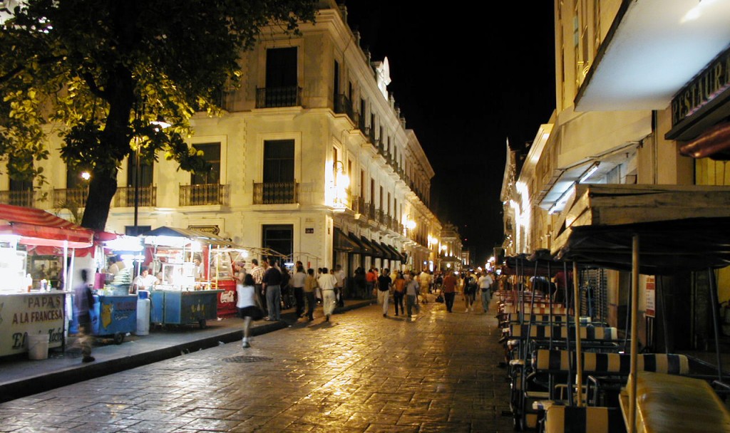 A colonial street at night bustling with shoppers and small market stalls.