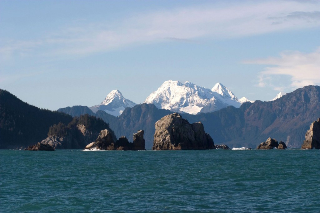 Large rocks jut out of the ocean with snow-capped mountains in the distance.