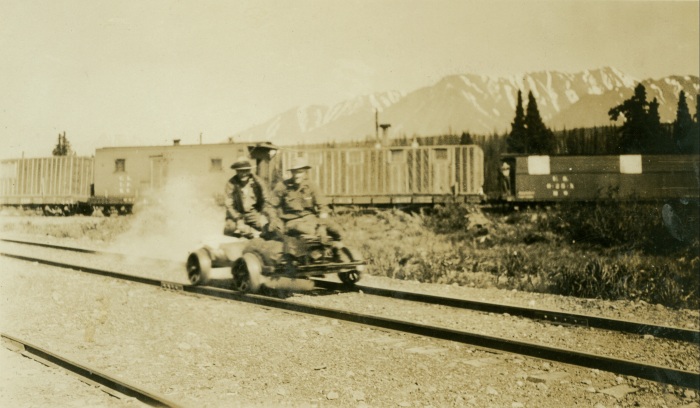 Two men on a four-wheeled velocipede roll down a railway with the mountains visible in the distance.