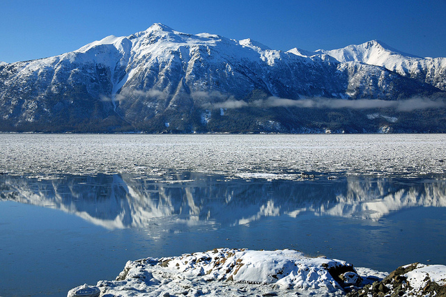 Snow-capped mountains reflect in water covered in melting ice.