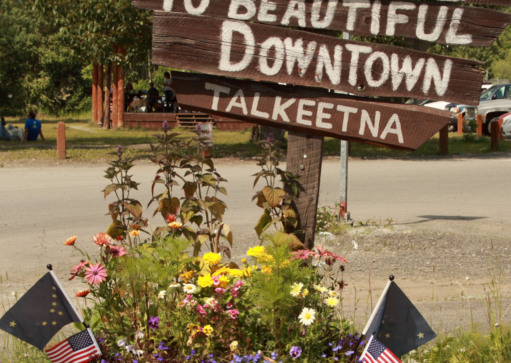Quirky hand-painted wooden sign reads Welcome to Beautiful Downtown Talkeetna and has flowers and american flags at the base.