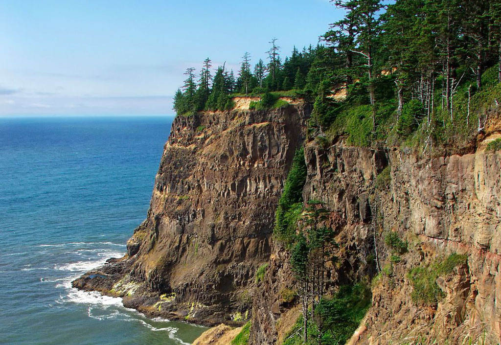 Waves crash at the base of an exposed cliff crowned with dense trees.