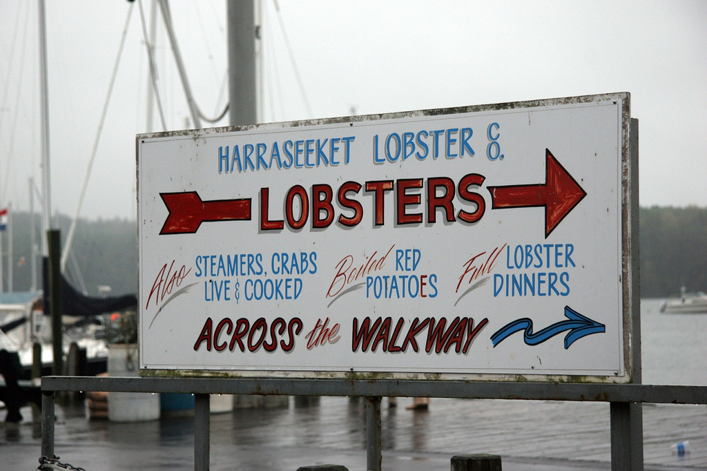 At the waterfront, a hand-lettered painted sign advertising the Harraseeket Lobster Co. and the food they serve.