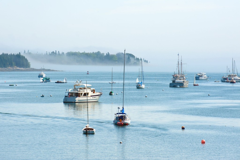 Yachts and boats move through the waters of Southwest Harbor while fog creeps along the coastline.