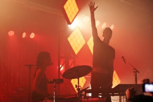 The duo Matt and Kim perform onstage in hazy red lighting.