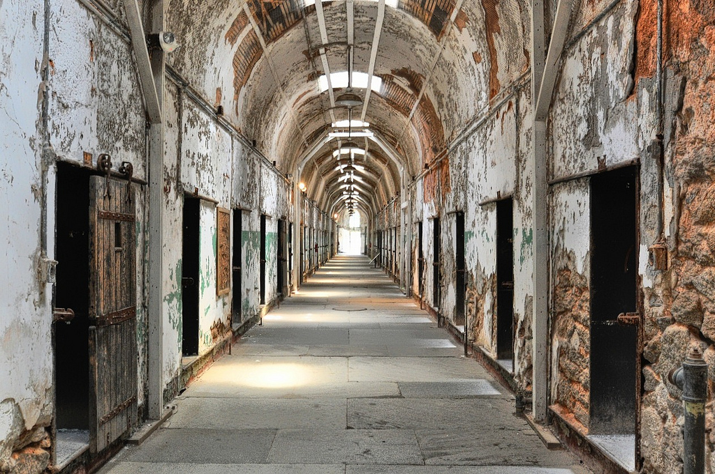 View down a dilapidated hallway with wooden cell doors and plaster peeling away from the stone walls.