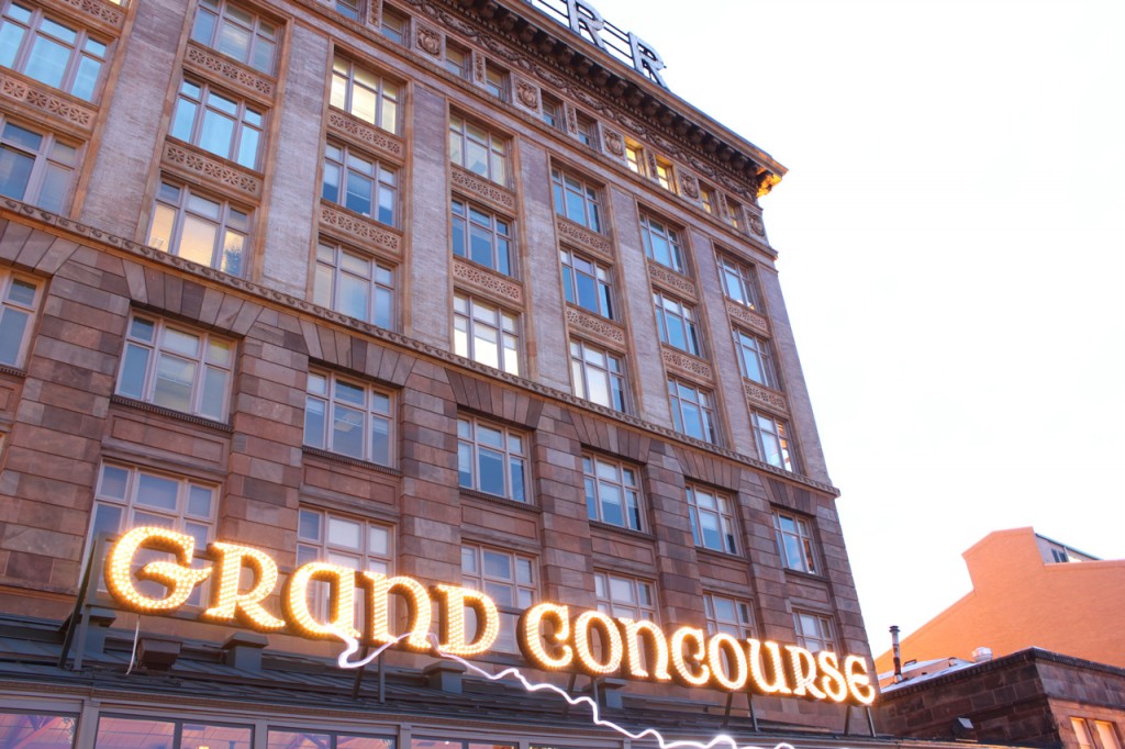 Grand Concourse is written in lights at the front of a windowed brick building.