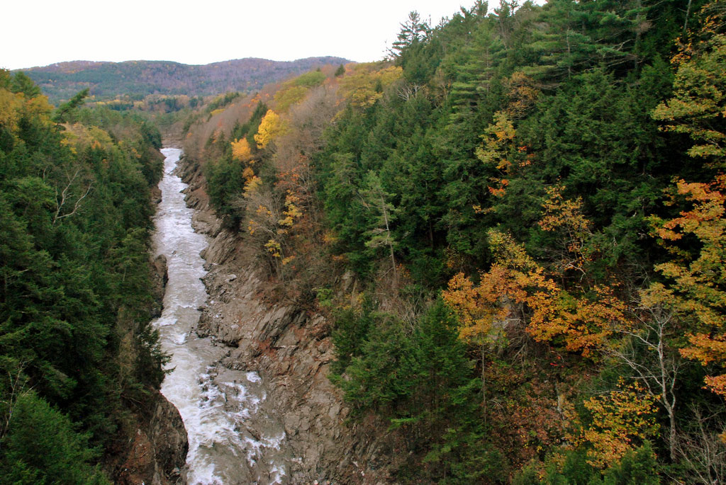 A narrow river rushes through a steep canyon lined with trees turning golden in Queechee.