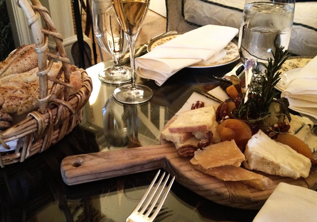 A selection of cheeses on a wooden board with rustic bread in a basket and wine glasses.