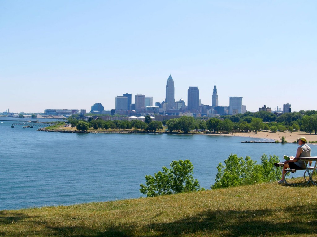 A person sits reading on a park bench on a clear day with the downtown skyline visible across the water.