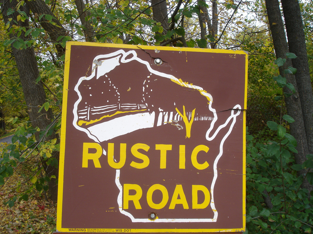 Green foliage just starting to turn is visible behind a road sign with an outline of the state and the text Rustic Road.