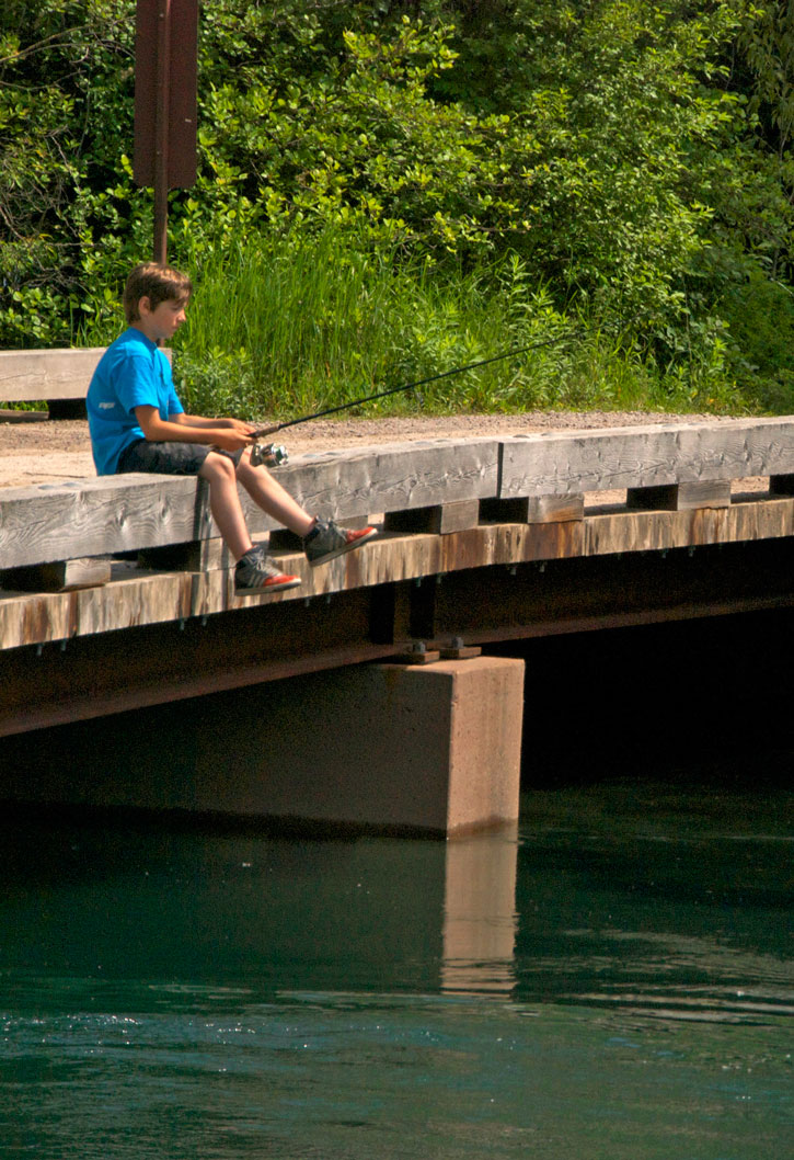 A boy sits on a footbridge fishing in the stream below.